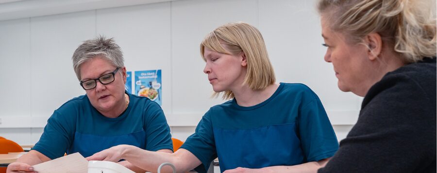 Mustatakkinen nainen keskustelee kahden hoitaja-asuihin pukeutuneen naisen kanssa.