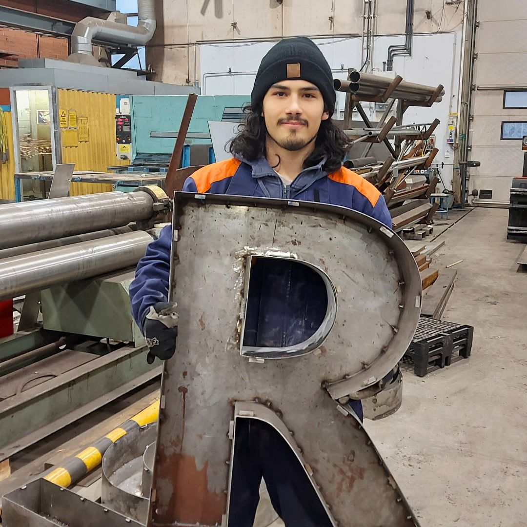 Metallialan opiskelija kädessään metallilevyitä tehty R-kirjain.
