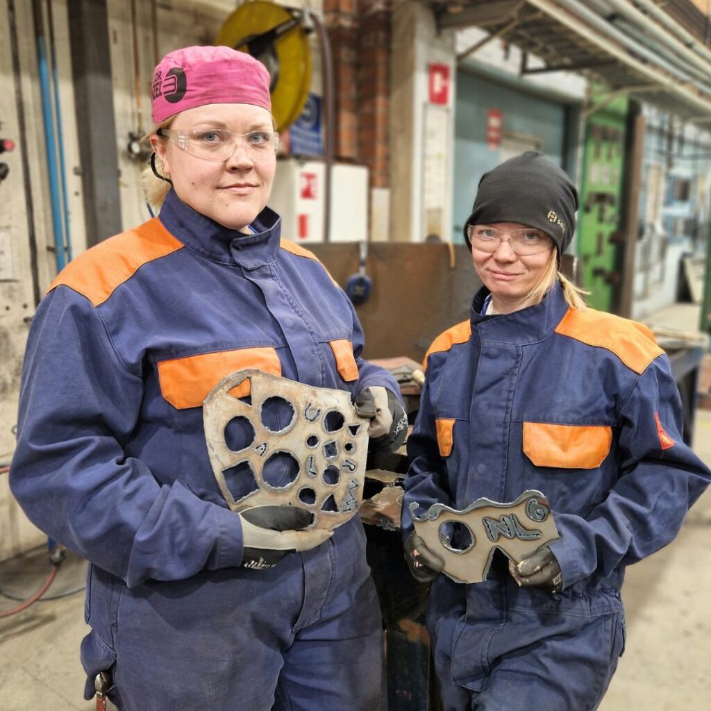 Kaksi metallialan naispuolista opiskelijaa seisoo metallityösalissa ja esittelee tekemiään harjoitustöitä.