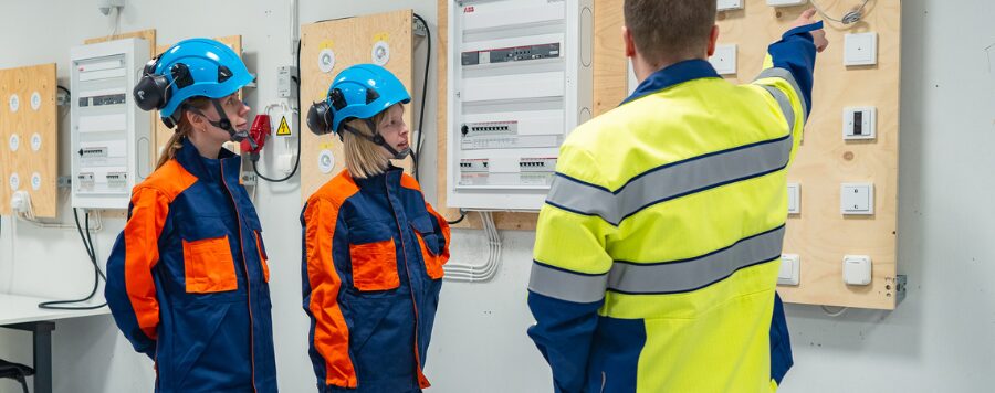 Kaksi sähkötyöntekijää seisoo vasemmalla kun työnjohtaja opastaa kuvan oikeassa reunassa.